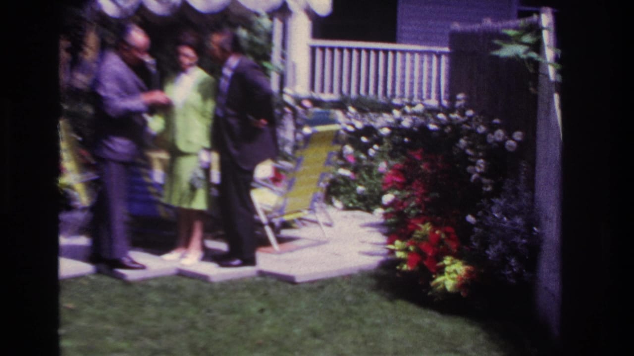 Adults stand in conversation beside a garden patio with flowers, chairs, and a shaded porch.
