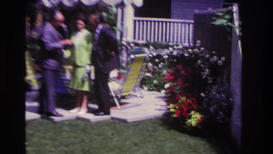 1967 Rockaway Beach 8mm frame with three adults standing by a backyard patio, yellow lounge chairs, flower beds, and porch railing.