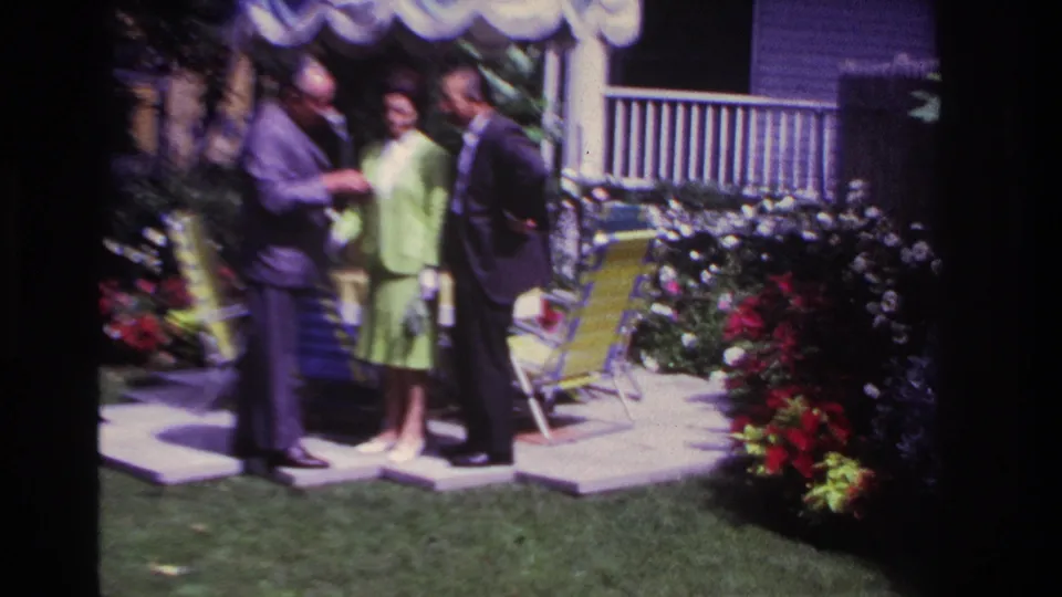 1967 Rockaway Beach backyard frame showing three adults on patio slabs, with lawn furniture, red flowers, and a white fence nearby.