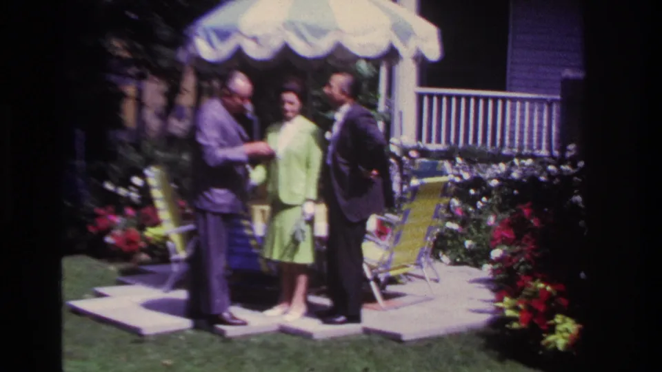 1967 Rockaway Beach home-movie frame of three adults gathered under a striped backyard umbrella beside chairs, flowers, and a porch.