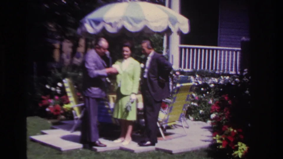 In 1967 Rockaway Beach home-movie footage, two suited men and a woman in a lime-green outfit stand together on a backyard patio.