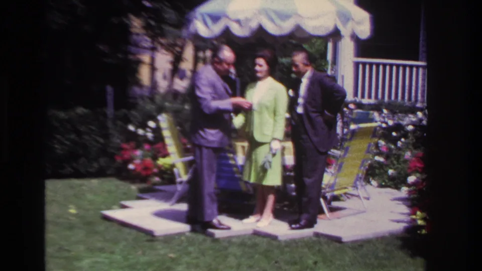 On the 1967 backyard patio, one suited man appears to show or hand something to the woman as another suited man watches nearby.