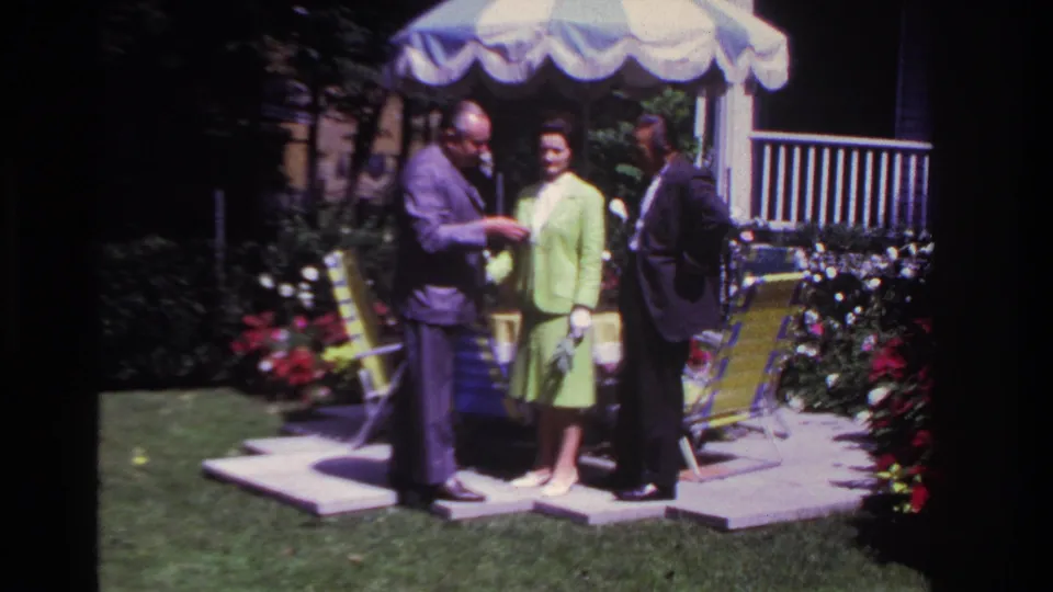 In 1967 Rockaway Beach home-movie footage, three adults stand in a sunny backyard as a man in a purple suit shows something near his wrist.