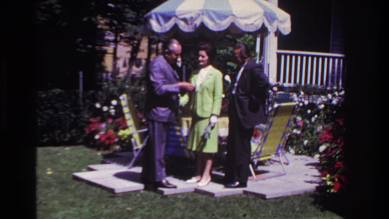 The backyard scene continues with the man in a purple suit gesturing toward his watch while a woman in green and a man in dark clothing watch.
