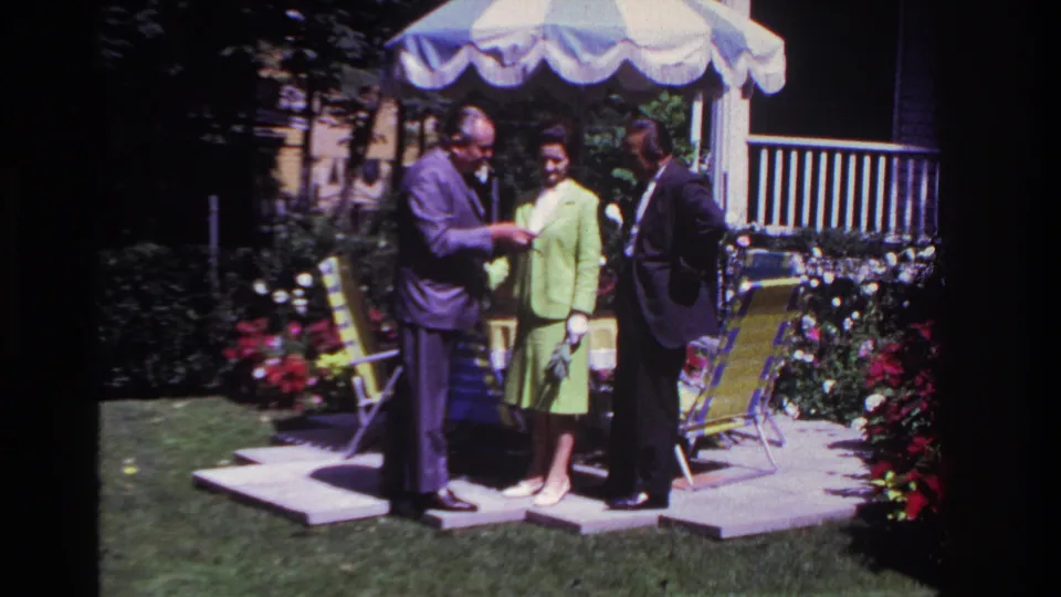 In the 1967 Rockaway Beach backyard, the older man appears to display his watch to a woman in a green suit and a man in a dark suit.