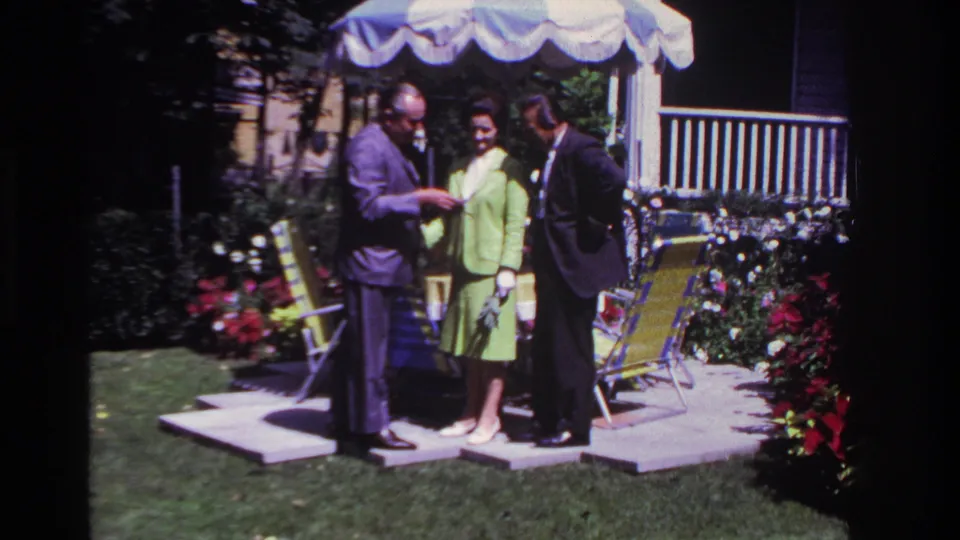 The clip ends with the group still gathered on the Rockaway Beach patio, framed by flowers, lawn chairs and a white-railed house exterior.