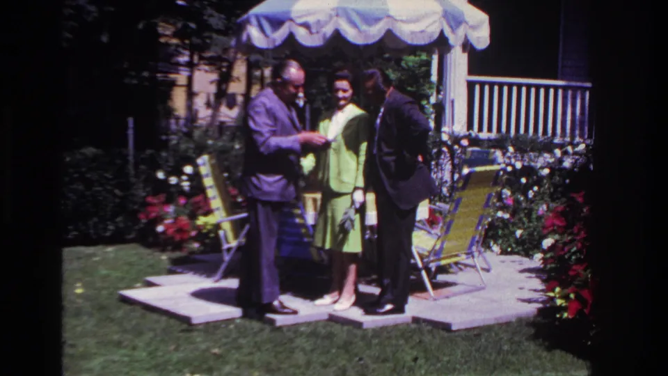 Backyard 8mm frame of three adults in formal clothing gathered beside flowers and folding chairs in 1967 Rockaway Beach.