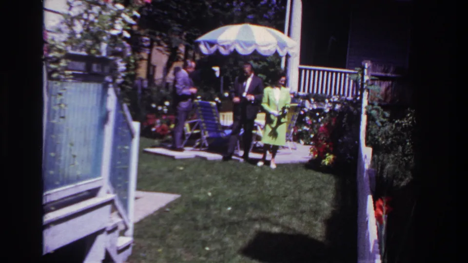 Wider 1967 Rockaway Beach backyard view with three adults on the patio, a lawn, flowers, and a white railing.