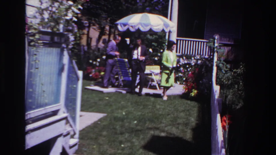 1967 Rockaway Beach home-movie frame showing adults in dress clothing in a backyard with a striped umbrella, flowers, and an open white gate.
