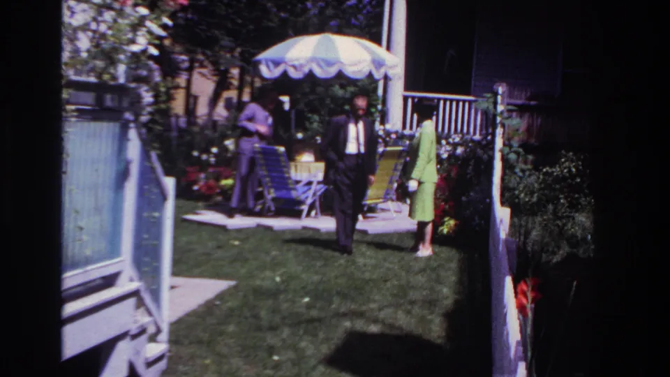 1967 Rockaway Beach home-movie still showing a man in a dark suit crossing a lawn, with a woman and another man near patio chairs.