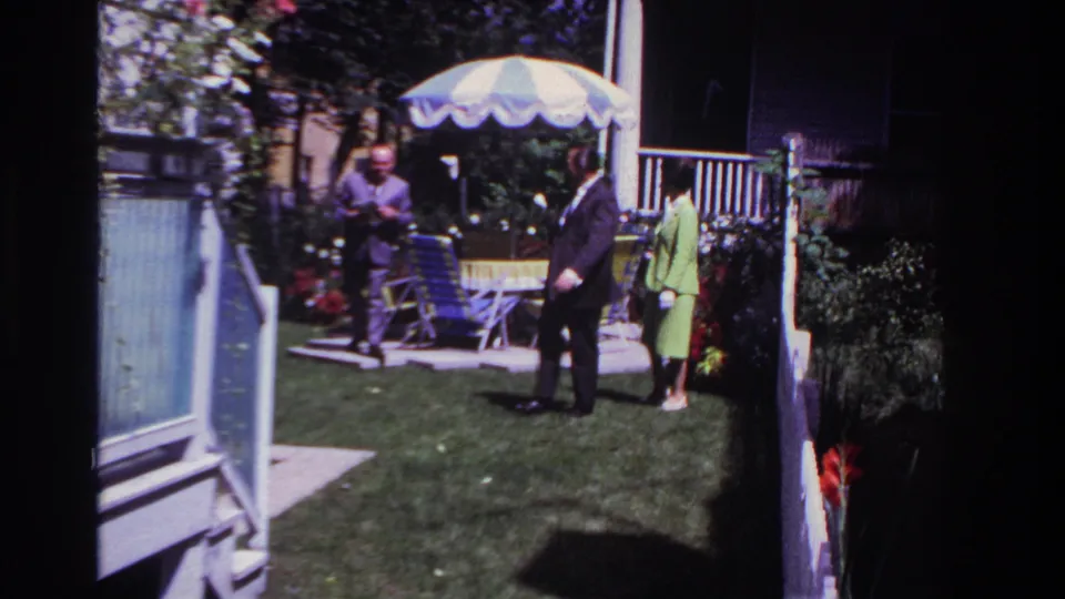 1967 Rockaway Beach frame showing three adults in a backyard beside folding chairs, flowers, porch railing, and a striped patio umbrella.