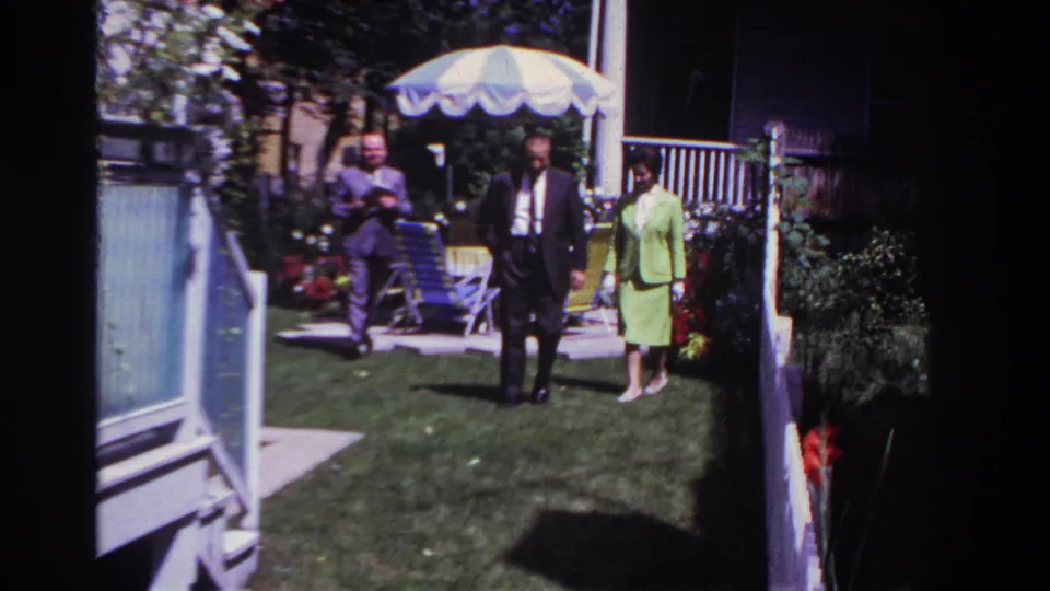 1967 Rockaway Beach home-movie frame showing three adults crossing a backyard lawn with a striped umbrella, flowers, and porch railings behind them.