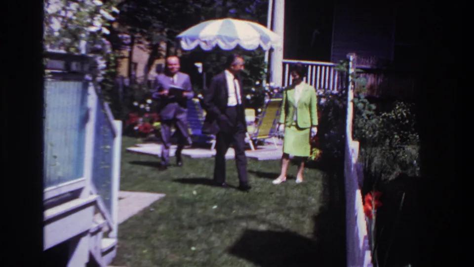 The group advances through a sunny Rockaway Beach backyard, with open garden gates, a striped umbrella, chairs, and bright flowers visible.