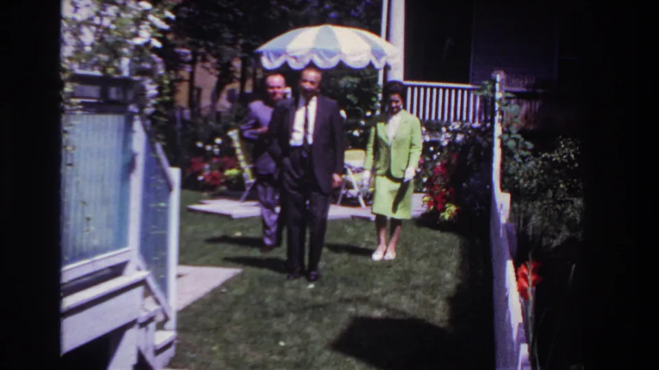 A suited man walks nearest the camera while two adults follow across a sunny residential lawn in the 1967 Rockaway Beach home-movie scene.