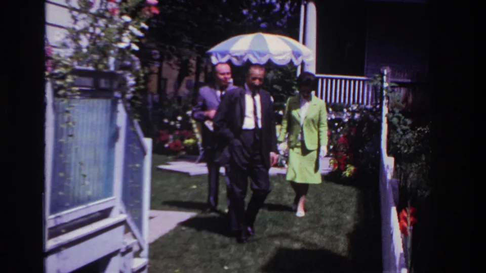 The adults near the camera at the end of the short backyard sequence, framed by flowers, fence posts, porch railings, and a striped patio umbrella.