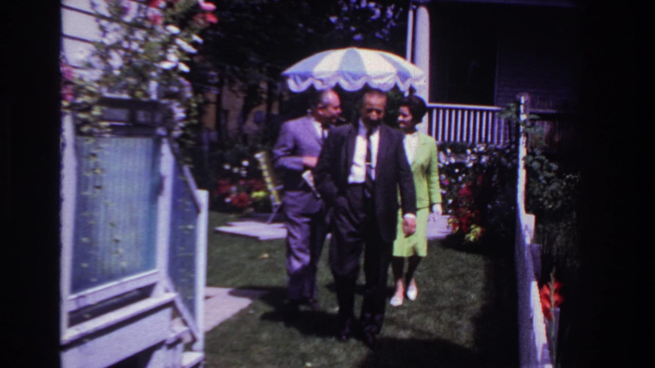 In 1967 Rockaway Beach home-movie footage, three formally dressed adults walk through a backyard near flowers, a porch, and a white fence.