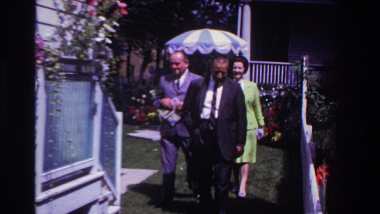 A man in a dark suit leads two dressed adults through a Rockaway Beach backyard, with a parasol, porch, flowers, and lawn visible.