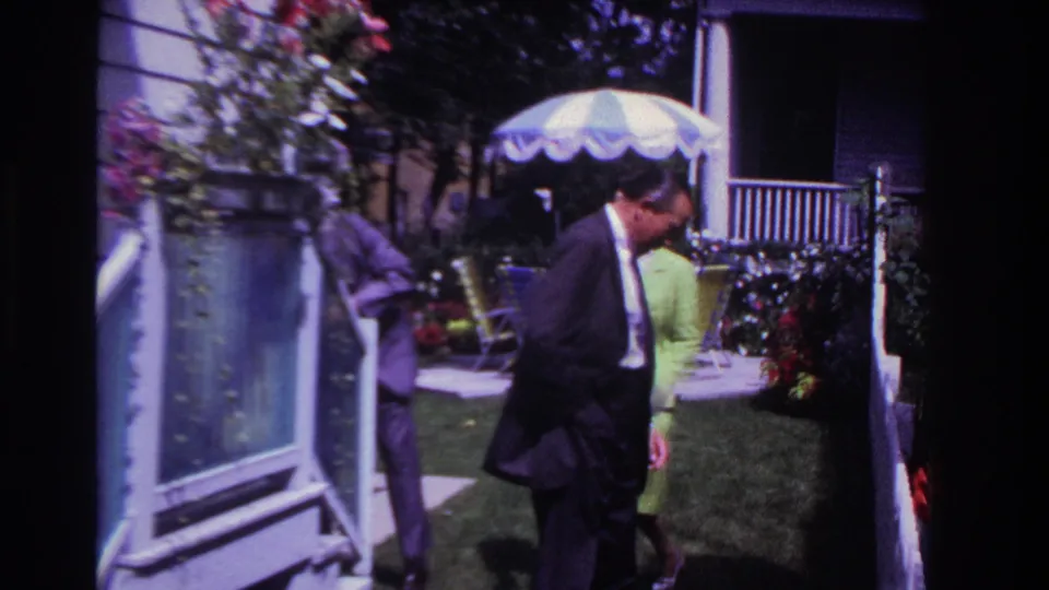 The dark-suited man moves past the camera in the 1967 backyard scene as the woman in green and another suited adult remain near the parasol.
