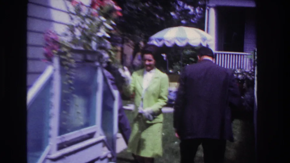 At the end of the Rockaway Beach home-movie clip, a woman in lime green stands near the white fence while a suited man faces away.