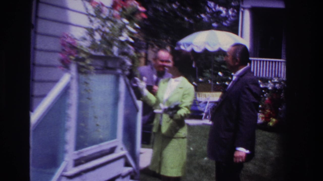 1967 Rockaway Beach home-movie frame showing a woman in a green suit and white gloves beside two suited men outside a house.
