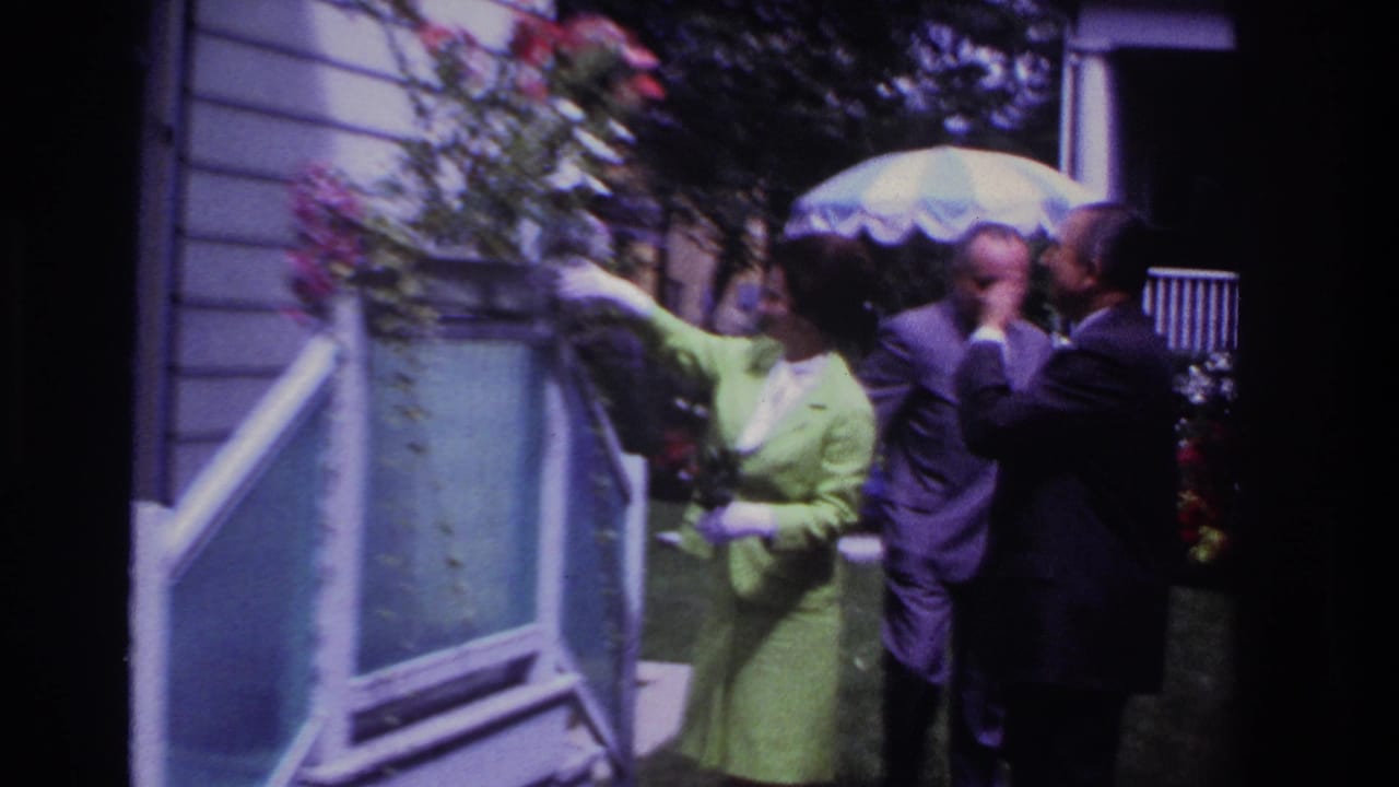 Woman in a green suit points toward flowers or the house while two men stand under an umbrella.