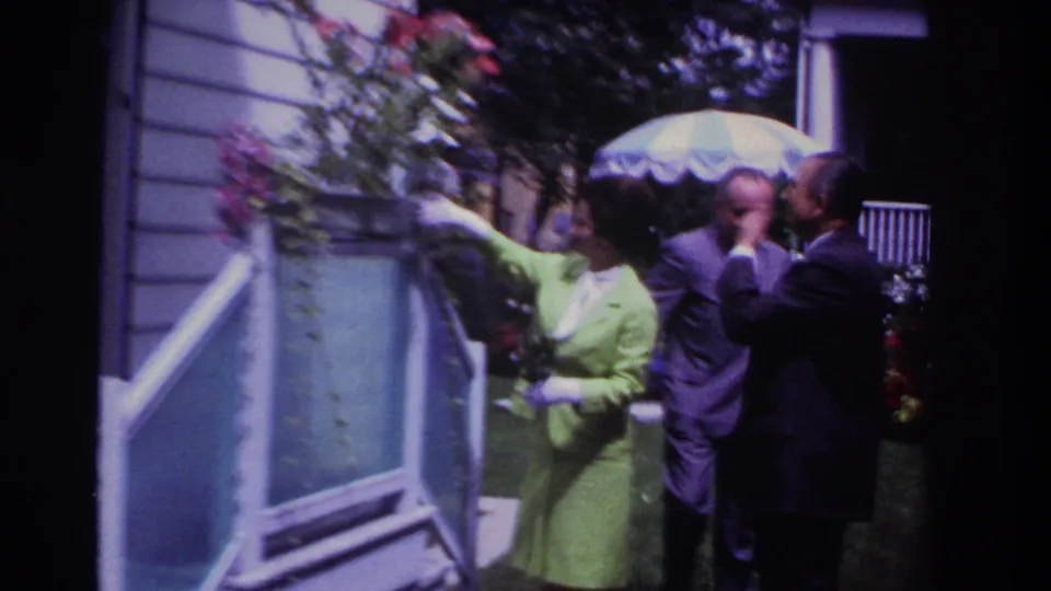 1967 Rockaway Beach frame with a woman gesturing outside a white house as two suited men confer near a patterned patio umbrella.