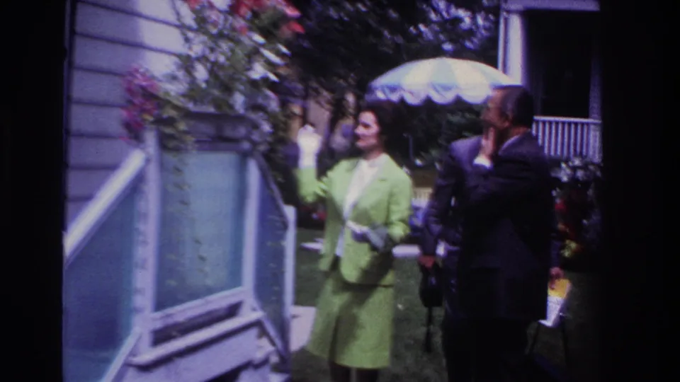 1967 Rockaway Beach home-movie frame showing a woman in green and two suited men outside a residence with flowers and lawn visible.
