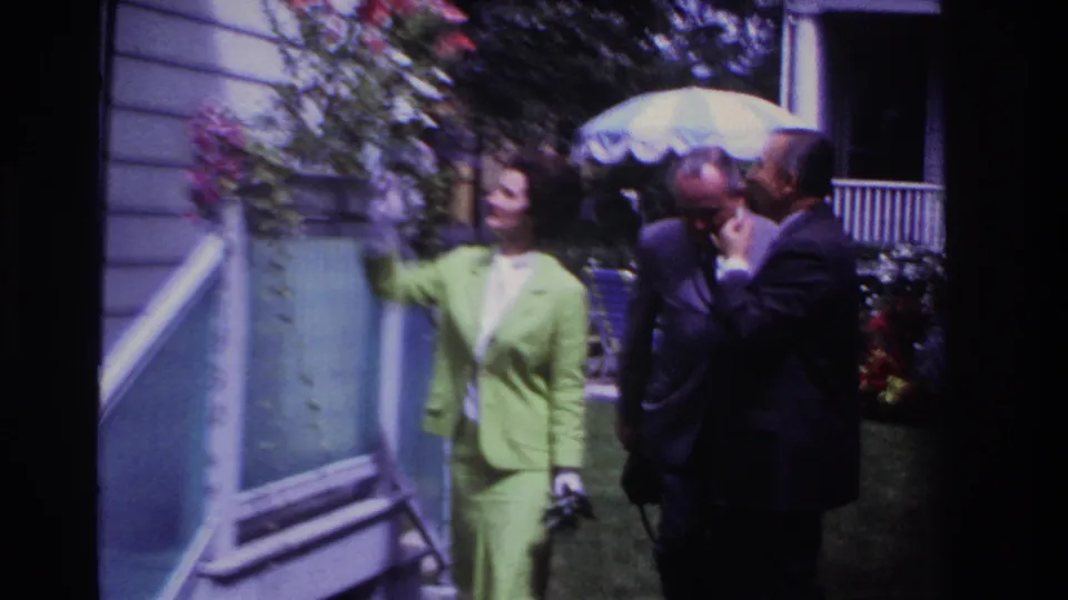 1967 Rockaway Beach frame of a woman in a bright green suit beside a house exterior, with two suited men and flowers behind her.