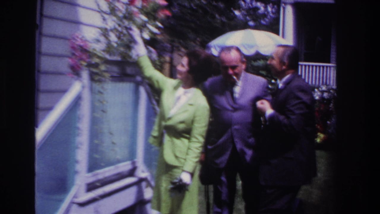 Woman in a green suit reaches toward porch flowers while two suited men stand nearby.