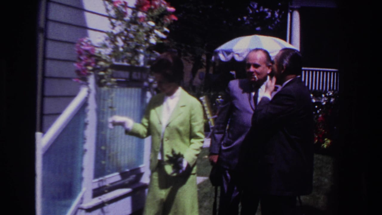 Woman stands beside a porch railing as two men in suits look on near a striped umbrella.