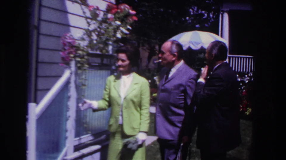 A smiling woman in white gloves stands by the porch while two men in suits face her near hanging flowers in the 1967 home-movie scene.