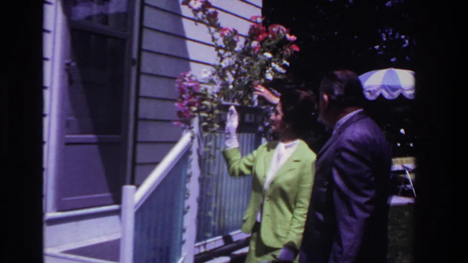 In 1967 Rockaway Beach home-movie footage, a woman in a lime green suit and white gloves gestures toward porch flowers beside a suited man.