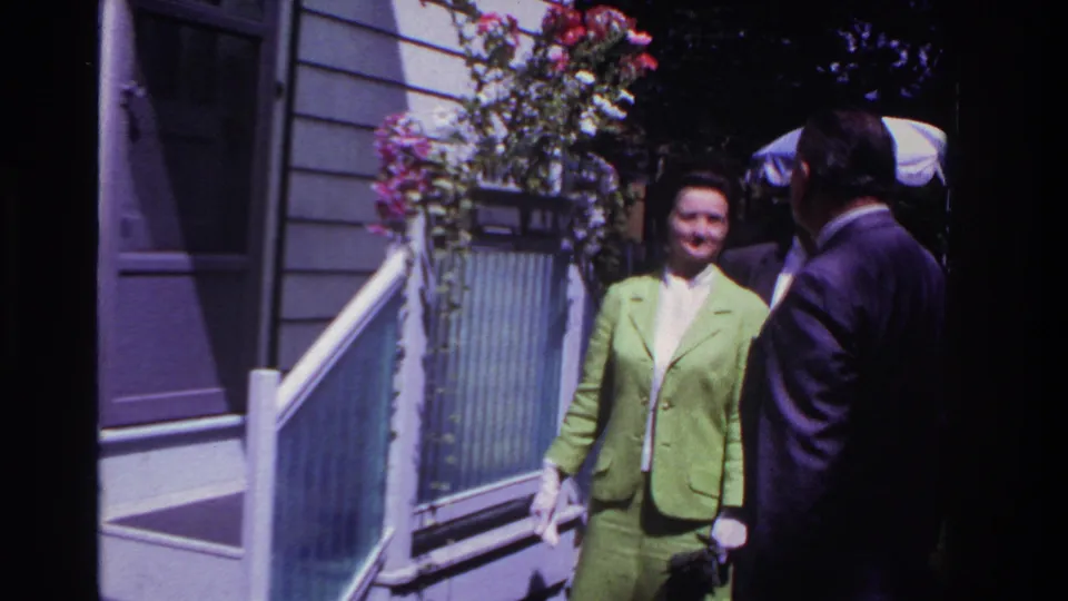 In the Rockaway Beach 1967 clip, the woman smiles on porch steps while two men in dark suits stand nearby, one partly in shadow.