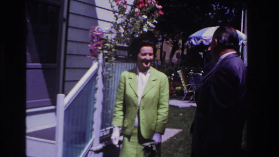 The woman in the lime green suit faces the camera in 1967 Rockaway Beach footage, standing near porch railings and climbing flowers.