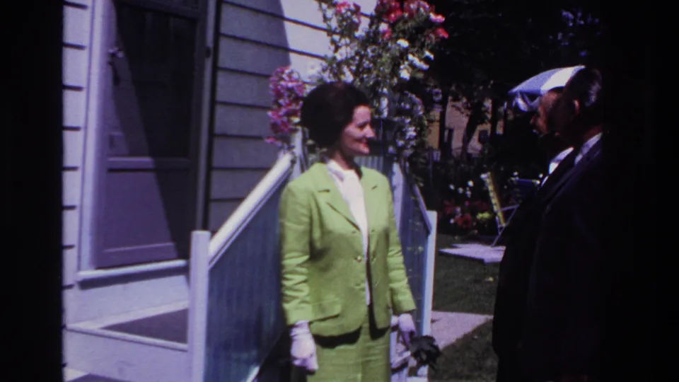At the end of the 1967 Rockaway Beach clip, the woman turns toward a suited man beside a porch with flowers and a shaded lawn behind them.