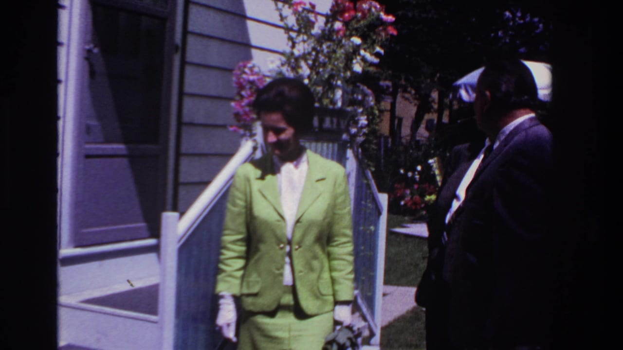 1967 Rockaway Beach frame with the woman in a lime green suit near a house entrance, partly framed by a man in a dark suit.