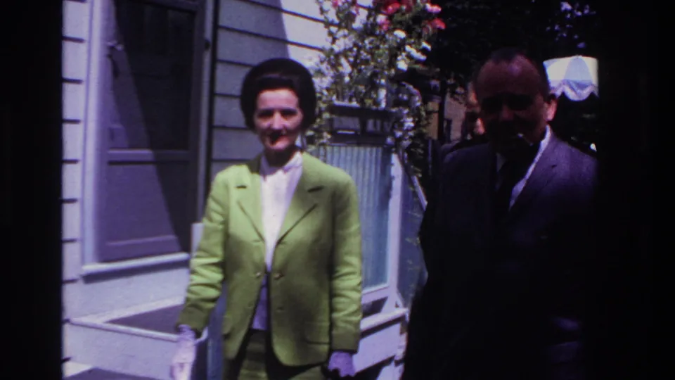 Rockaway Beach 1967 home-movie frame showing a smiling woman in a lime green suit and a man in a dark suit outside a house.