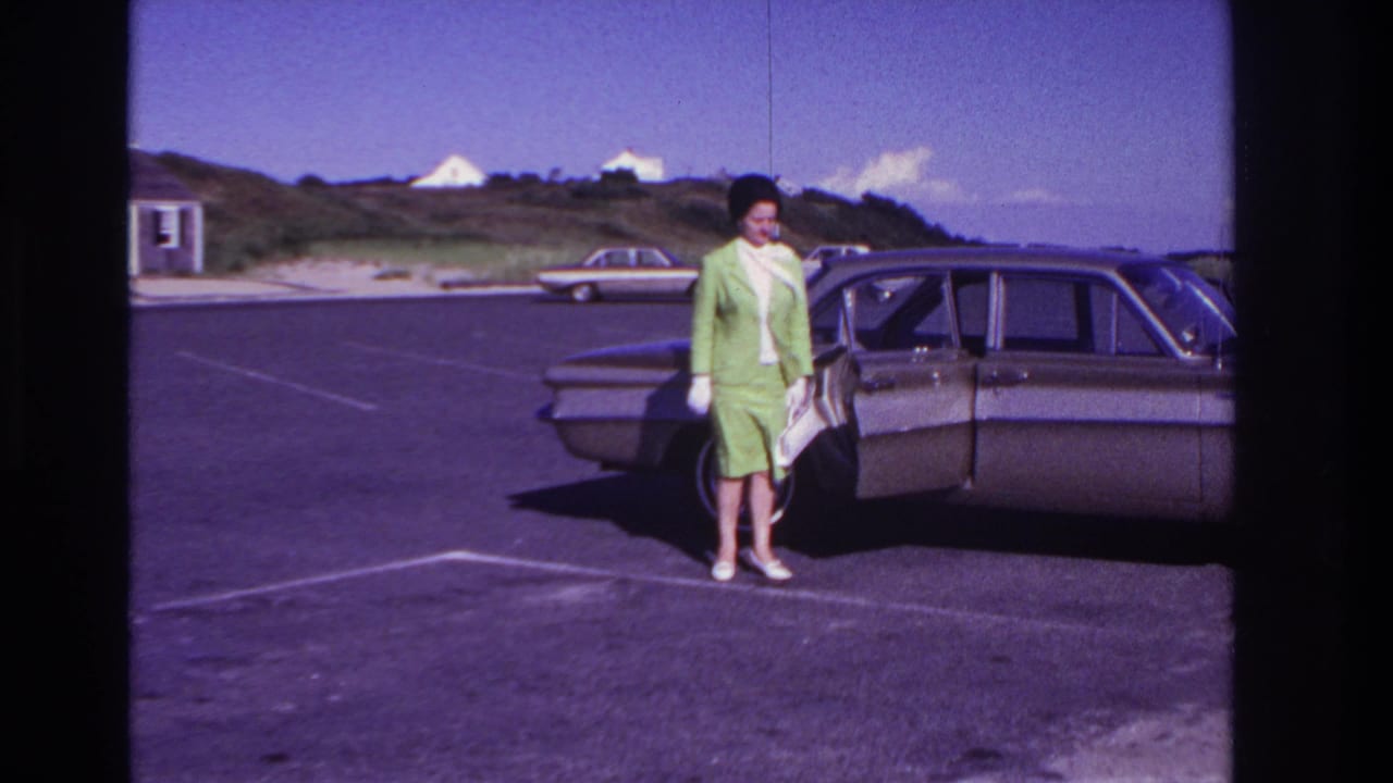 At Rockaway Beach in 1967, a woman in a green suit and white gloves stands beside a brown 1960s sedan in a sunny parking lot.
