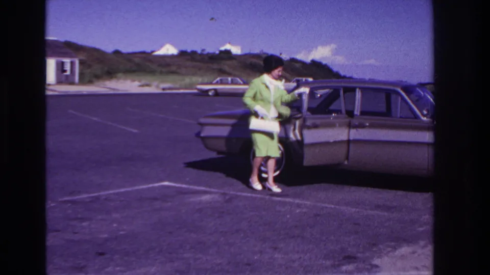 In the 1967 Rockaway Beach parking lot, the woman turns toward the sedan door while holding her purse and standing on painted asphalt lines.