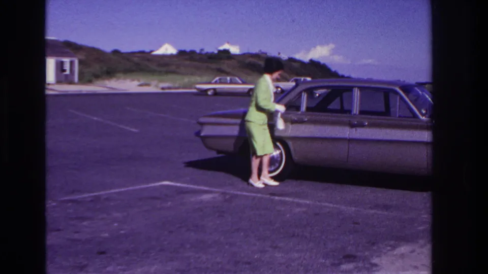 The woman stands close to the brown sedan door at Rockaway Beach, with the beachside lot, dunes and distant parked car still visible.