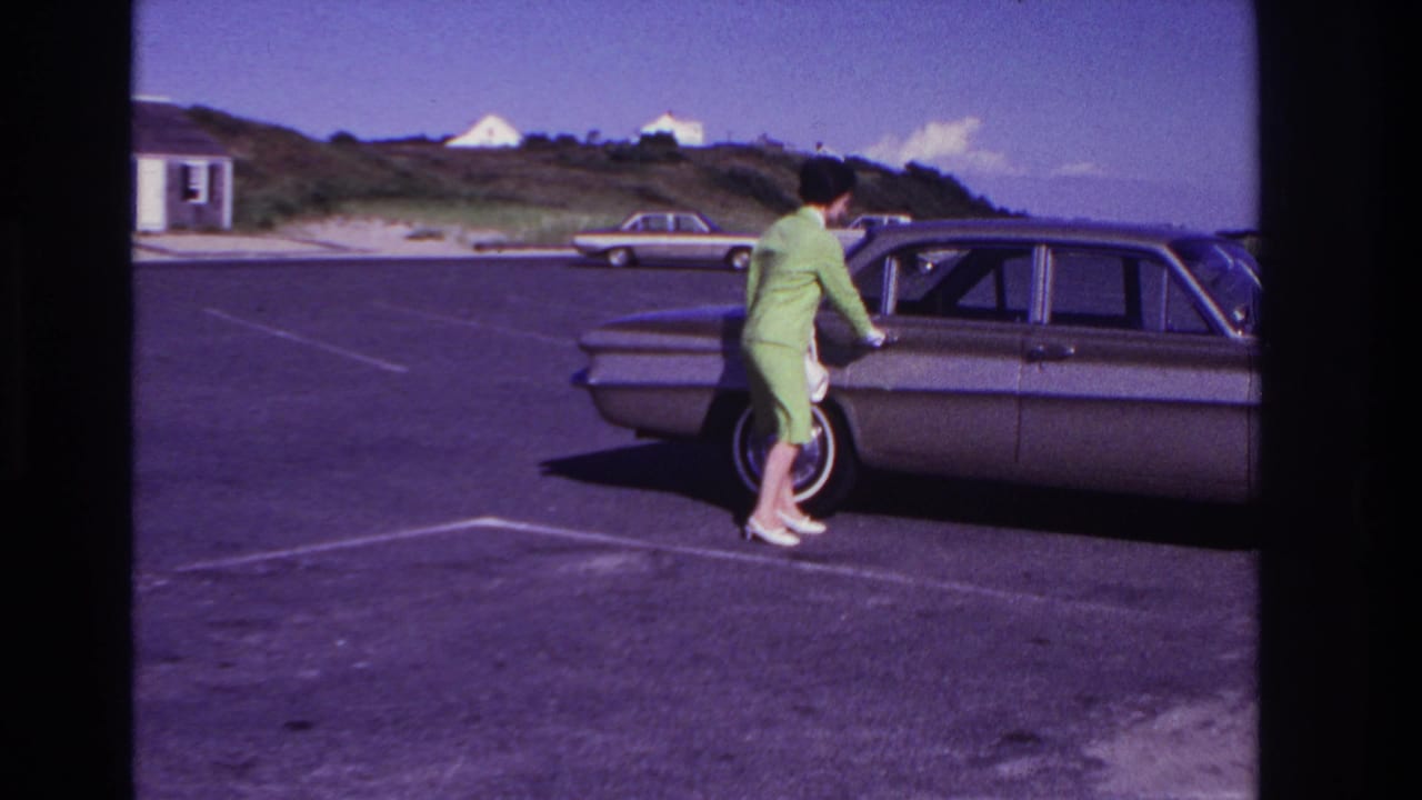 At Rockaway Beach in 1967, a woman in a green suit and white gloves stands by an open sedan door in a sunny parking lot.