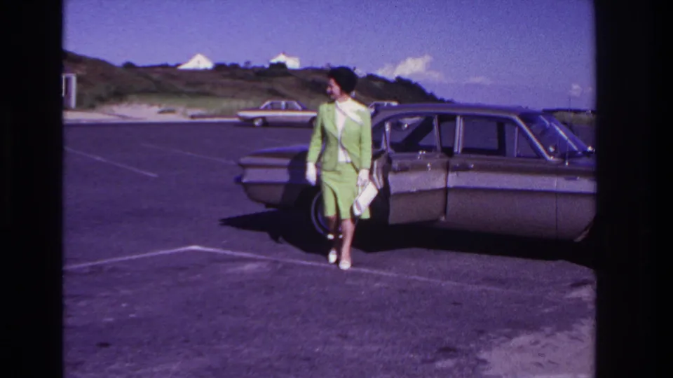 At Rockaway Beach in 1967, the woman walks from the sedan with her white purse, framed by a paved lot, dunes, and distant houses.