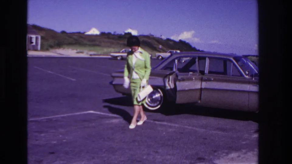 At Rockaway Beach in 1967, the woman pauses near the sedan's open door with her purse as sunlight falls across the parking lot.