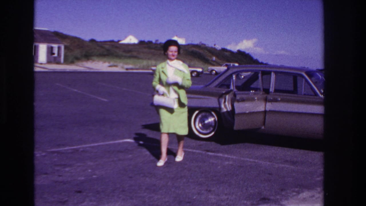 In 1967 Rockaway Beach footage, a woman in a green suit, white gloves, and heels stands near a classic sedan in a parking lot.