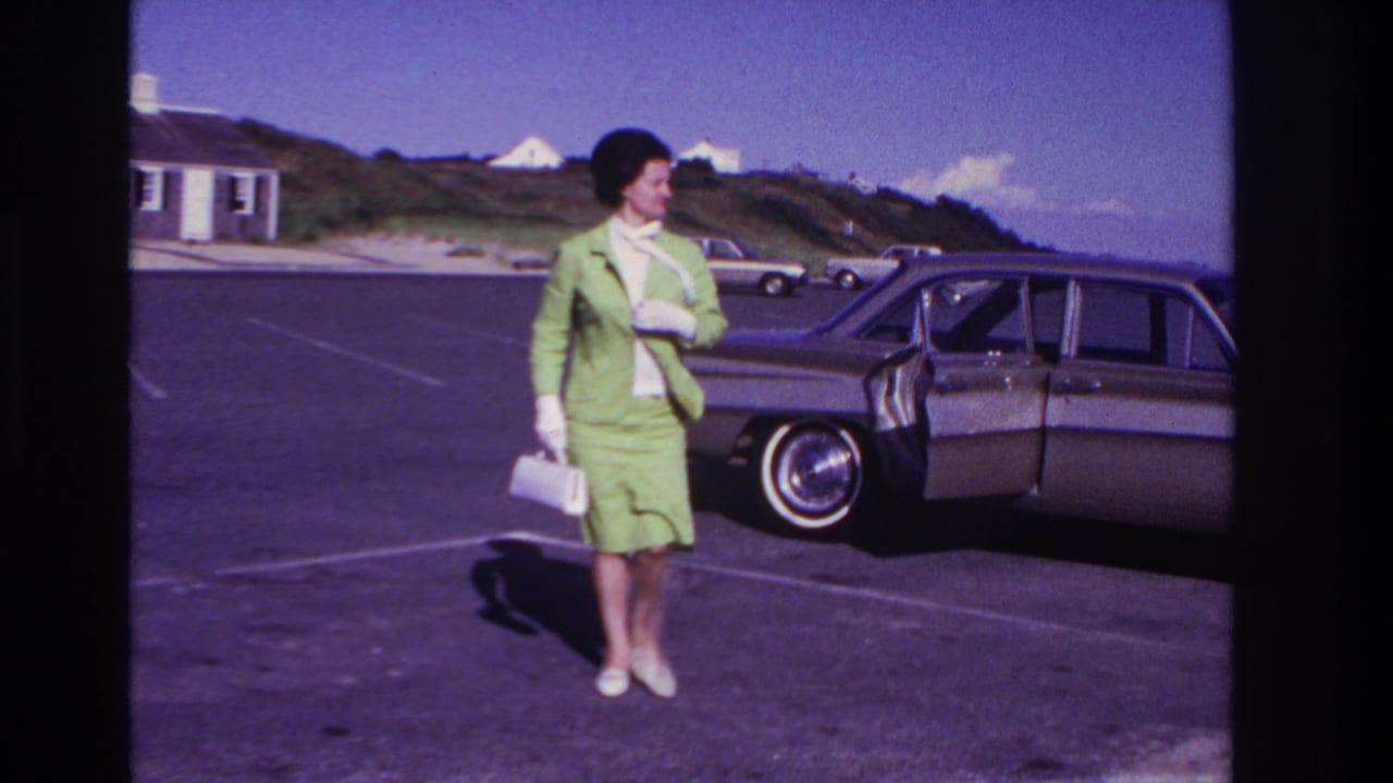 Woman in green suit turns beside the open area near a parked sedan.