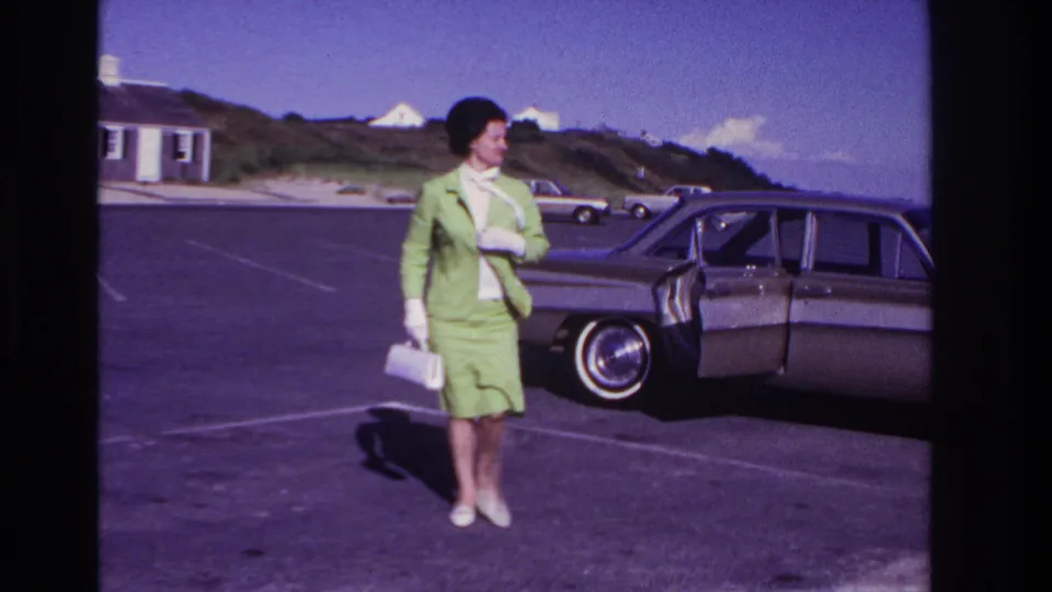 The woman in a green suit and white gloves turns away from the sedan, with beach dunes and small buildings behind her.