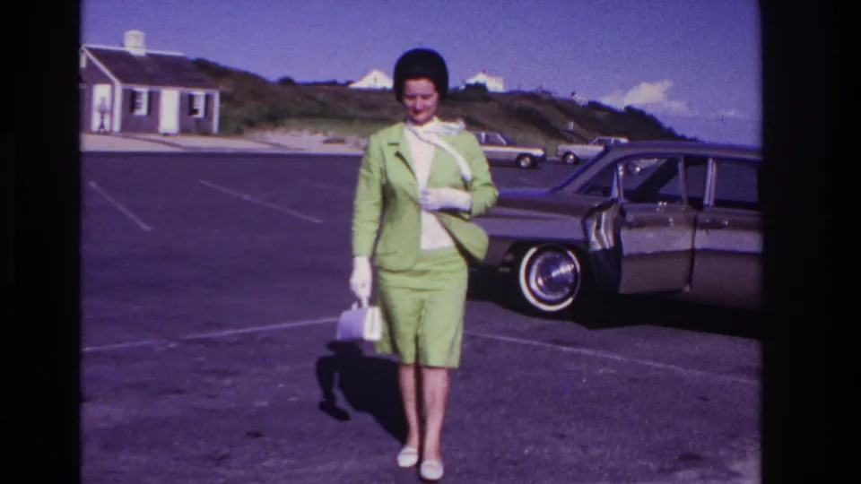 A woman in 1960s dress walks toward the camera at Rockaway Beach, carrying a white purse near a classic sedan.