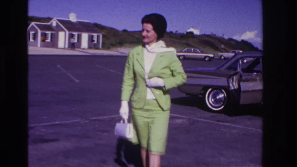 The woman smiles in a green skirt suit and white accessories, framed beside a classic sedan in 1967 Rockaway Beach home-movie footage.