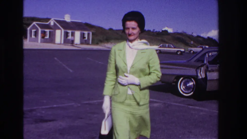 At Rockaway Beach in the stated 1967 footage, a woman in a green suit and white gloves stands near a parked mid-century car.