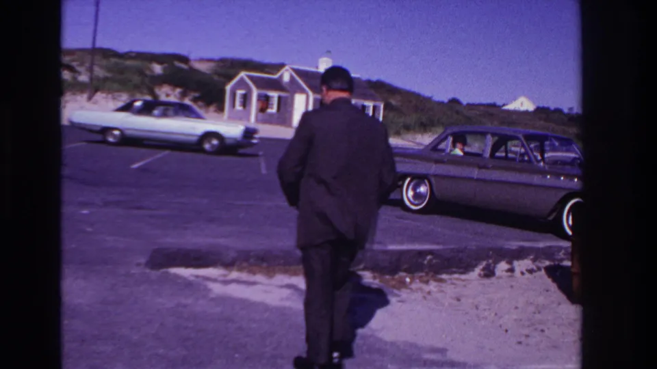 The suited man walks away from the camera across a paved beach parking lot, with classic cars and low coastal buildings visible.